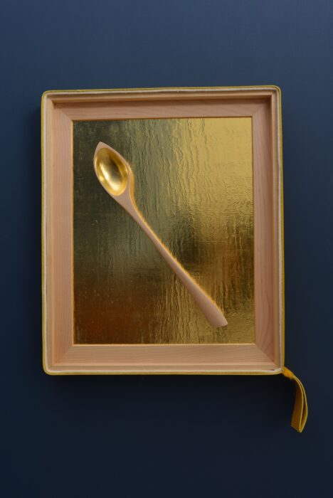 Hand-carved birch spoon resting against a textured gold leaf background, framed in birch with leather and felt accents.