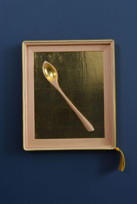 Hand-carved birch spoon resting against a textured gold leaf background, framed in birch with leather and felt accents.