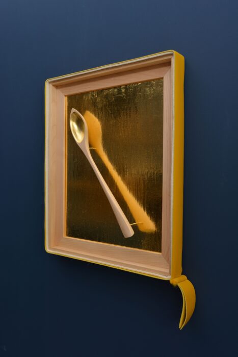 Hand-carved birch spoon resting against a textured gold leaf background, framed in birch with leather and felt accents.